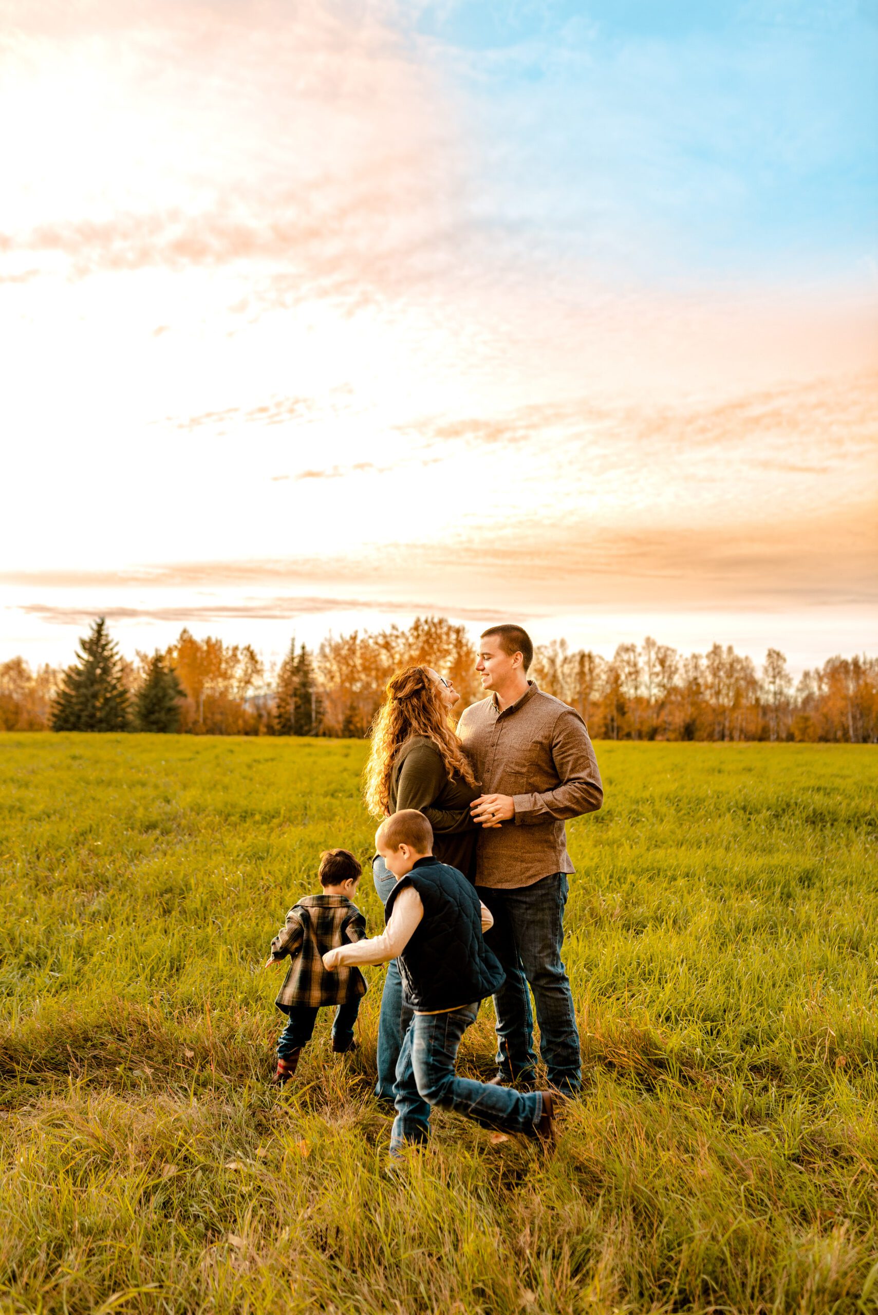 A couple is holding hands during their family session as their two boys run around each other during the late Fall season in an open field.
