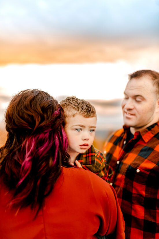 a mom holds and embraces her son with her back turned toward the camera as her son looks off into the distance, her husband looking at both of them with a smile from Fayetteville, NC family photographer 